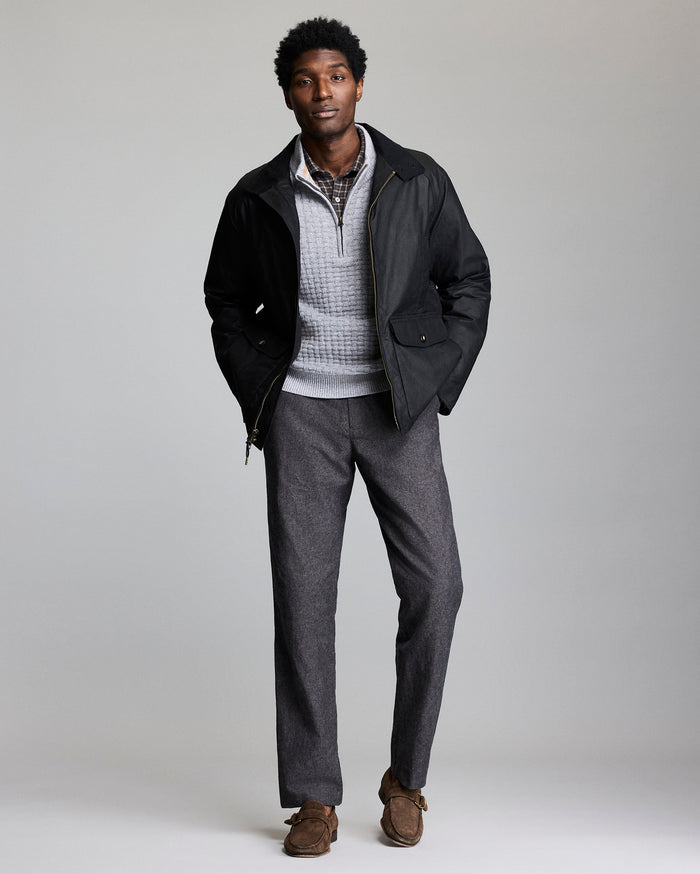 A man stands against a plain background wearing a black jacket over a light gray quarter-zip sweater, a checked shirt, gray trousers, and brown loafers. His hands are in his jacket pockets and he is looking at the camera.