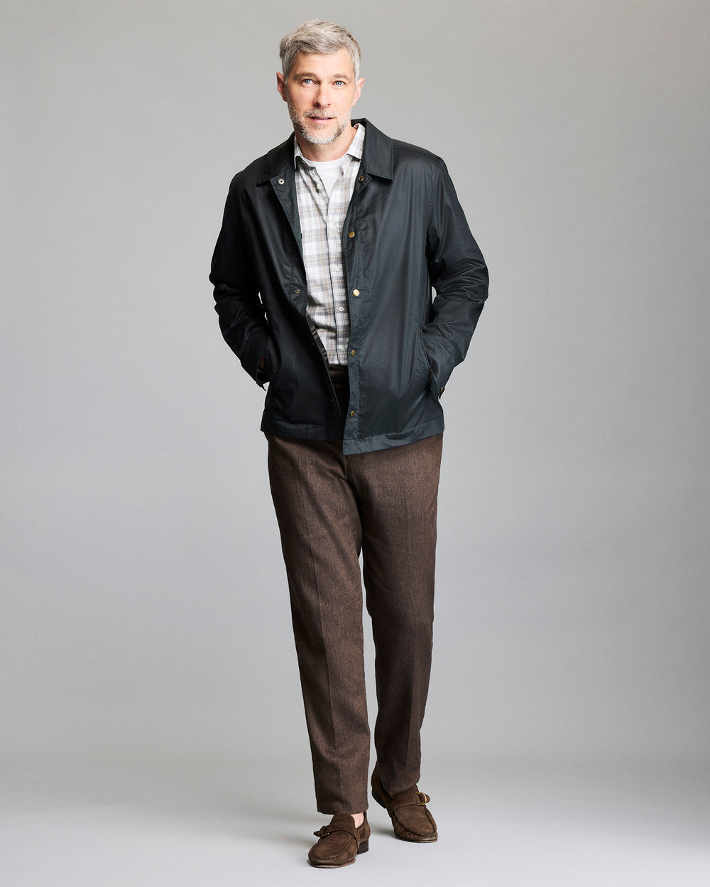 A man with gray hair and a beard is standing against a plain background, wearing a black jacket over a light plaid shirt, brown pants, and brown suede shoes, with his hands in his jacket pockets.