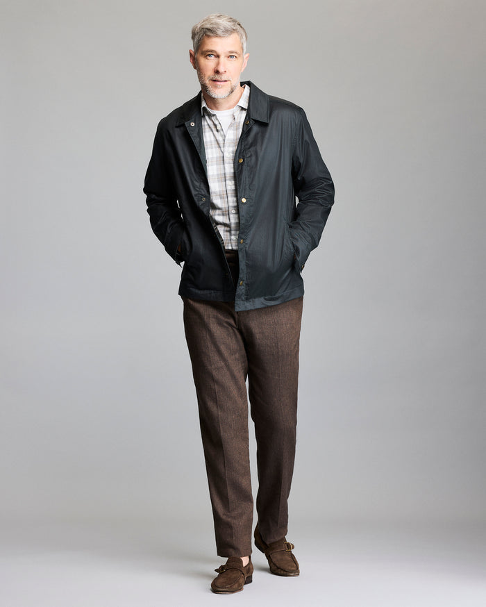 A man with gray hair and a beard is standing against a plain background, wearing a black jacket over a light plaid shirt, brown pants, and brown suede shoes, with his hands in his jacket pockets.