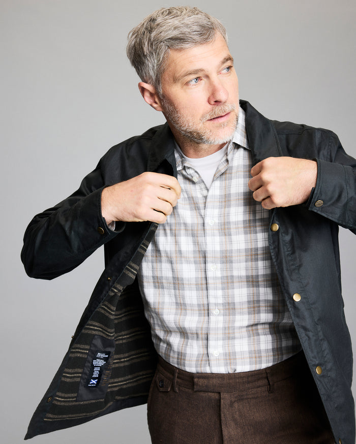 A man with short gray hair and a beard wears a checked shirt and brown pants while opening a dark jacket, looking off to the side against a plain gray background.