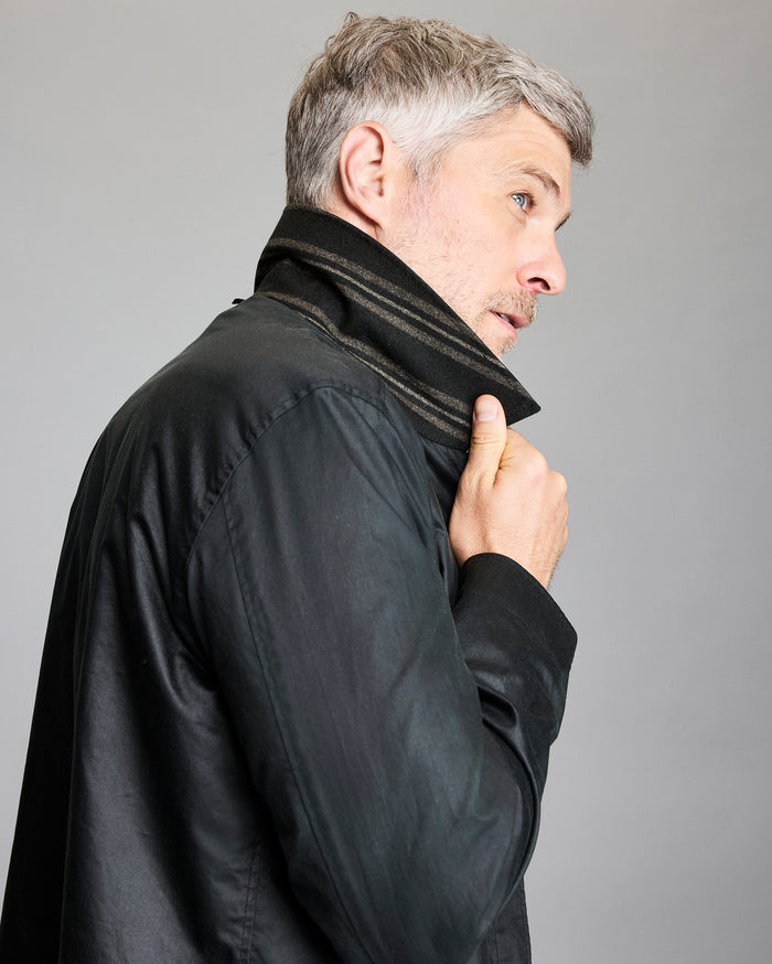 A man with short gray hair and stubble lifts the collar of his black jacket, looking off to the side against a plain gray background.