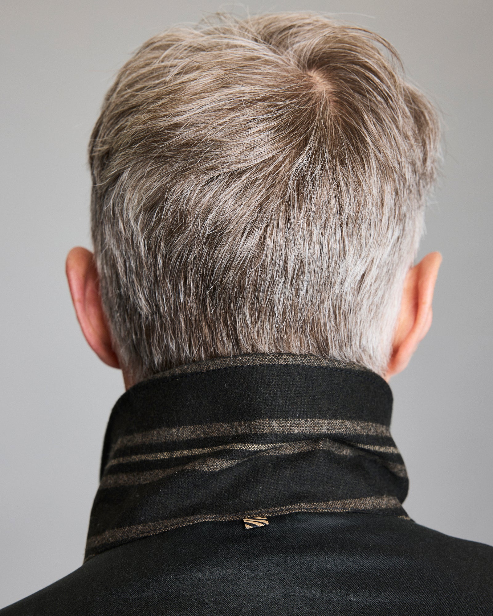 A person with short, gray hair is shown from behind, wearing a dark coat with the collar turned up. The background is plain and neutral.