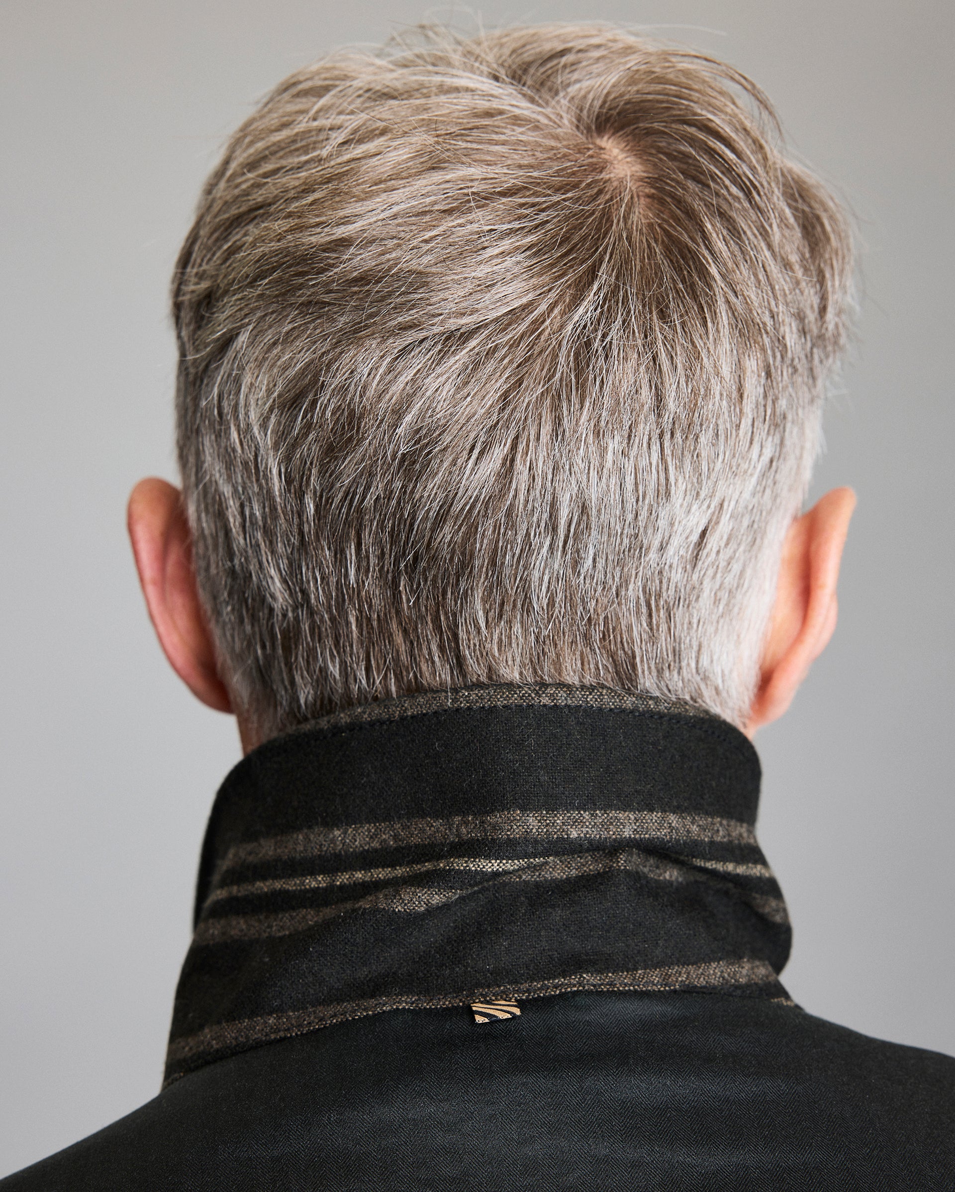 A person with short, gray hair is shown from behind, wearing a dark coat with the collar turned up. The background is plain and neutral.