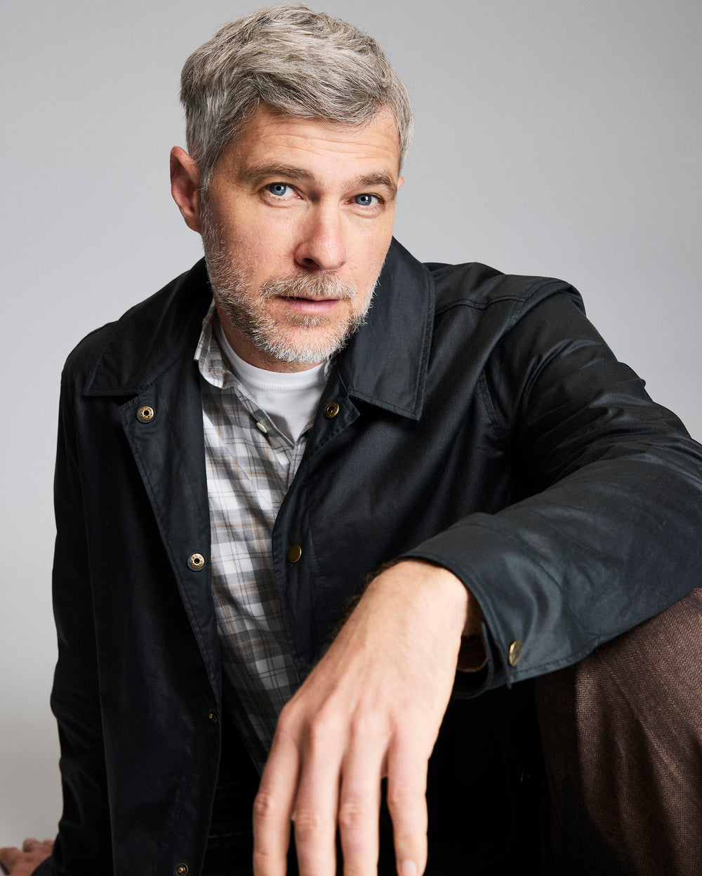 A man with short gray hair and a beard, wearing a black jacket over a plaid shirt, sits and looks directly at the camera with a neutral expression, resting his arm on his knee against a plain background.