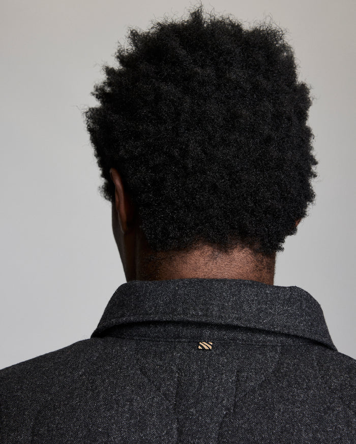 A person with short, curly black hair is shown from behind, wearing a dark textured coat with a small gold pin on the collar. The background is plain and light-colored.