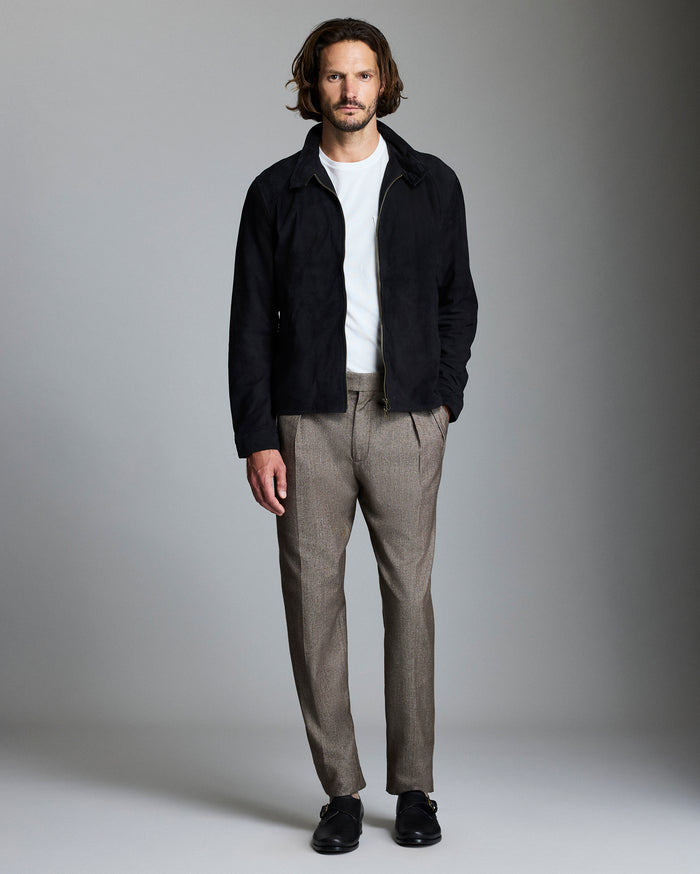 A man with wavy hair stands against a plain background, wearing a black jacket over a white shirt, brown checked trousers, and black shoes. One hand is in his pocket and he looks directly at the camera.