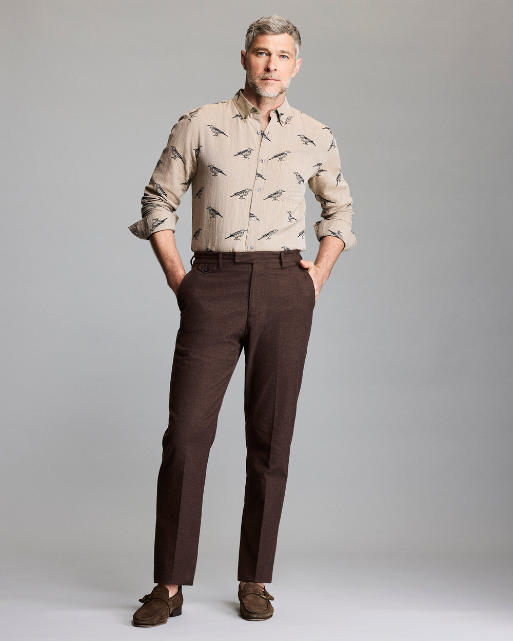 A man with gray hair and a beard stands against a plain background, wearing a beige button-up shirt with a bird print, dark brown dress pants, and brown loafers. His hands are in his pockets and he looks confidently ahead.