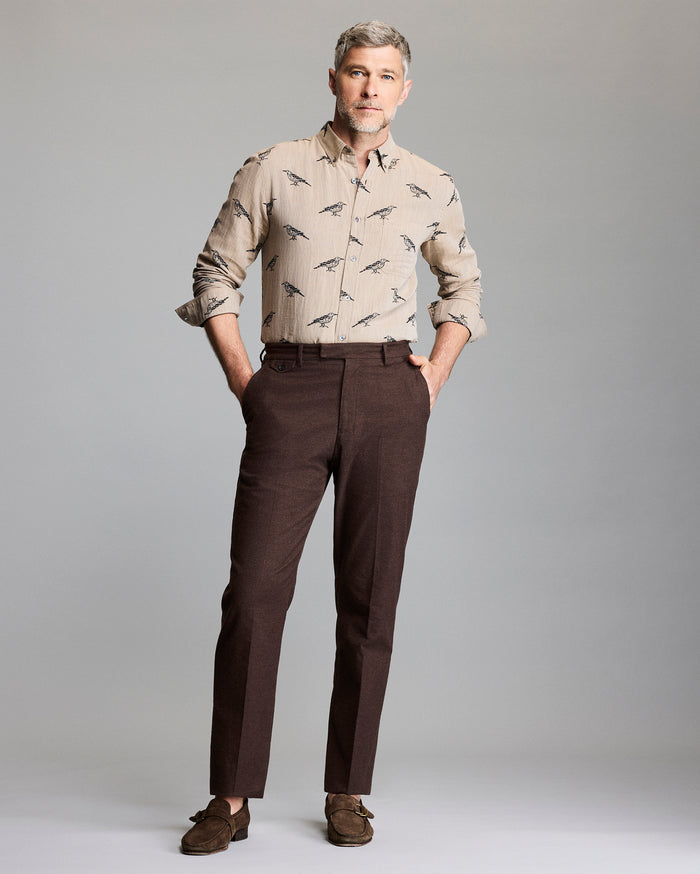 A man with gray hair and a beard stands against a plain background, wearing a beige button-up shirt with a bird print, dark brown dress pants, and brown loafers. His hands are in his pockets and he looks confidently ahead.