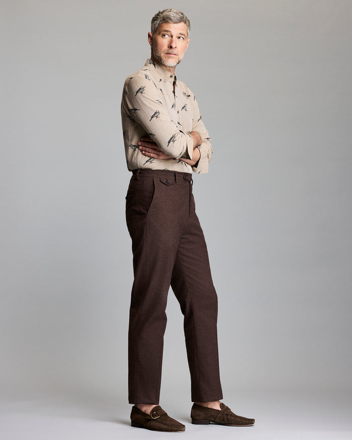 A man with short gray hair stands sideways with arms crossed, wearing a beige patterned button-up shirt, brown pants, and brown loafers, against a plain light gray background.