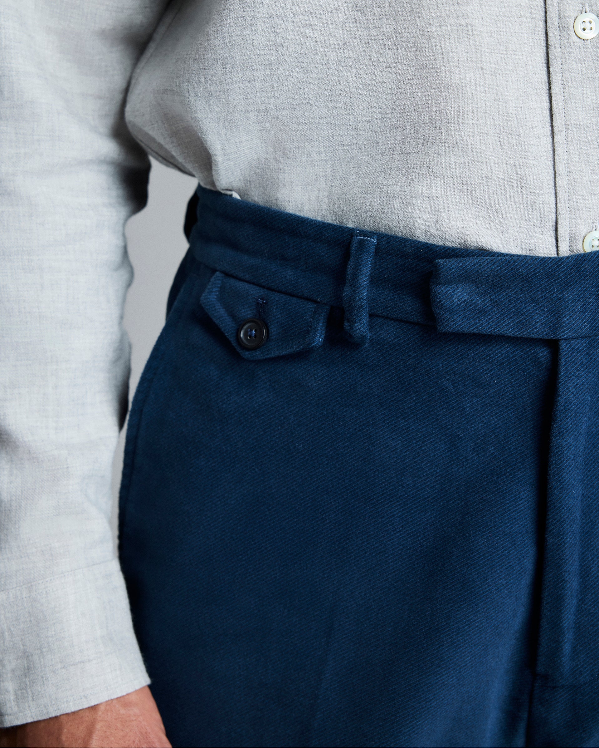 A person wearing a light grey button-up shirt tucked into high-waisted, dark blue pants with a belt and buttoned side-tab detail. Only the lower torso and part of one arm are visible.