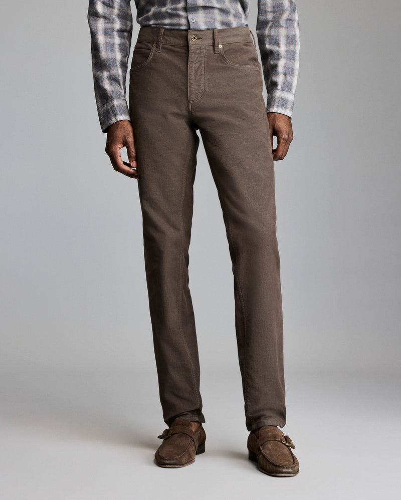 A person wearing brown straight-leg pants, a grey and white plaid shirt, and brown suede loafers stands against a plain light grey background. Only the lower half of the body is visible. Alternate