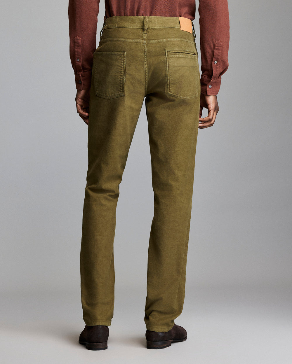 A person stands facing away, wearing olive green pants, a brown long-sleeve shirt, and dark brown shoes against a plain gray background.