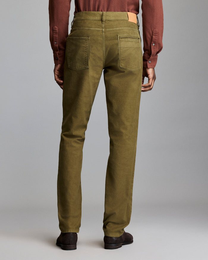 A person stands facing away, wearing olive green pants, a brown long-sleeve shirt, and dark brown shoes against a plain gray background.