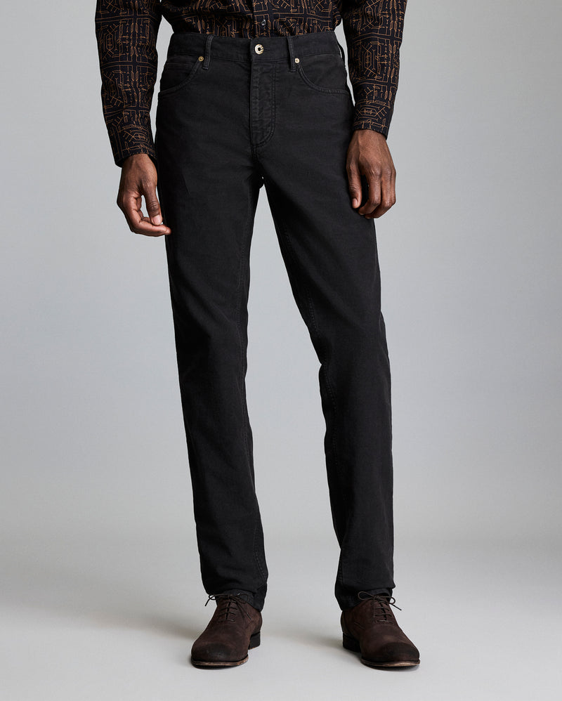 A man stands against a plain background wearing black pants, a patterned long-sleeve shirt, and dark brown dress shoes. His arms are relaxed at his sides, and his head is not visible in the frame. Alternate