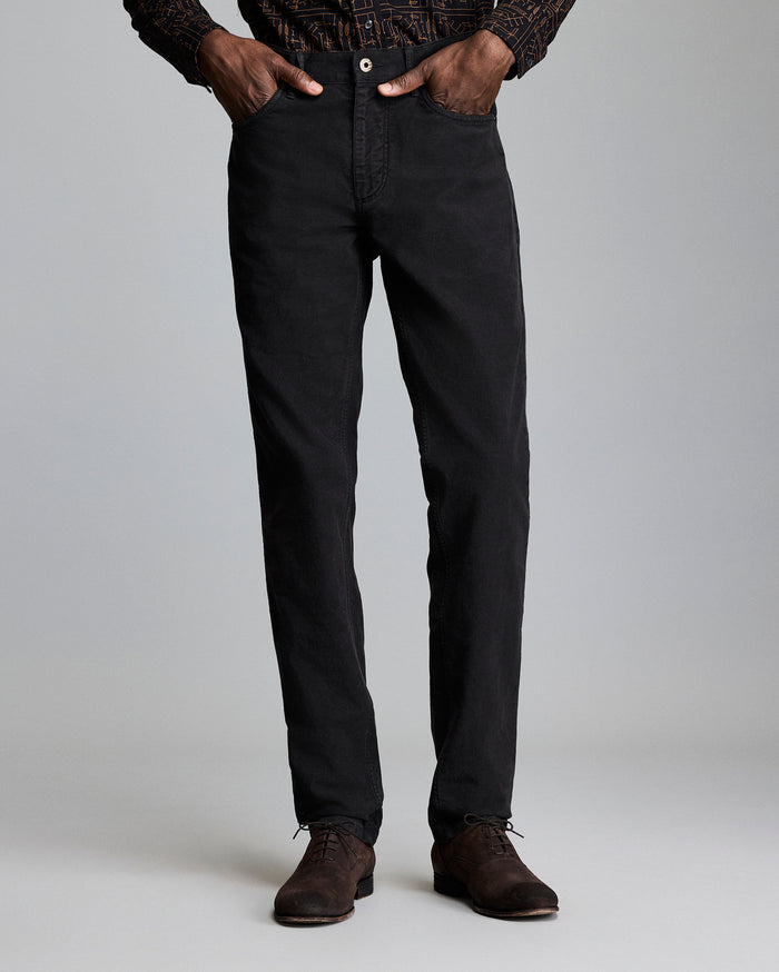 A person stands against a plain background, wearing black straight-leg pants, a patterned long-sleeve shirt, and brown dress shoes. Their hands are in their pockets, and their upper body is partially out of frame.