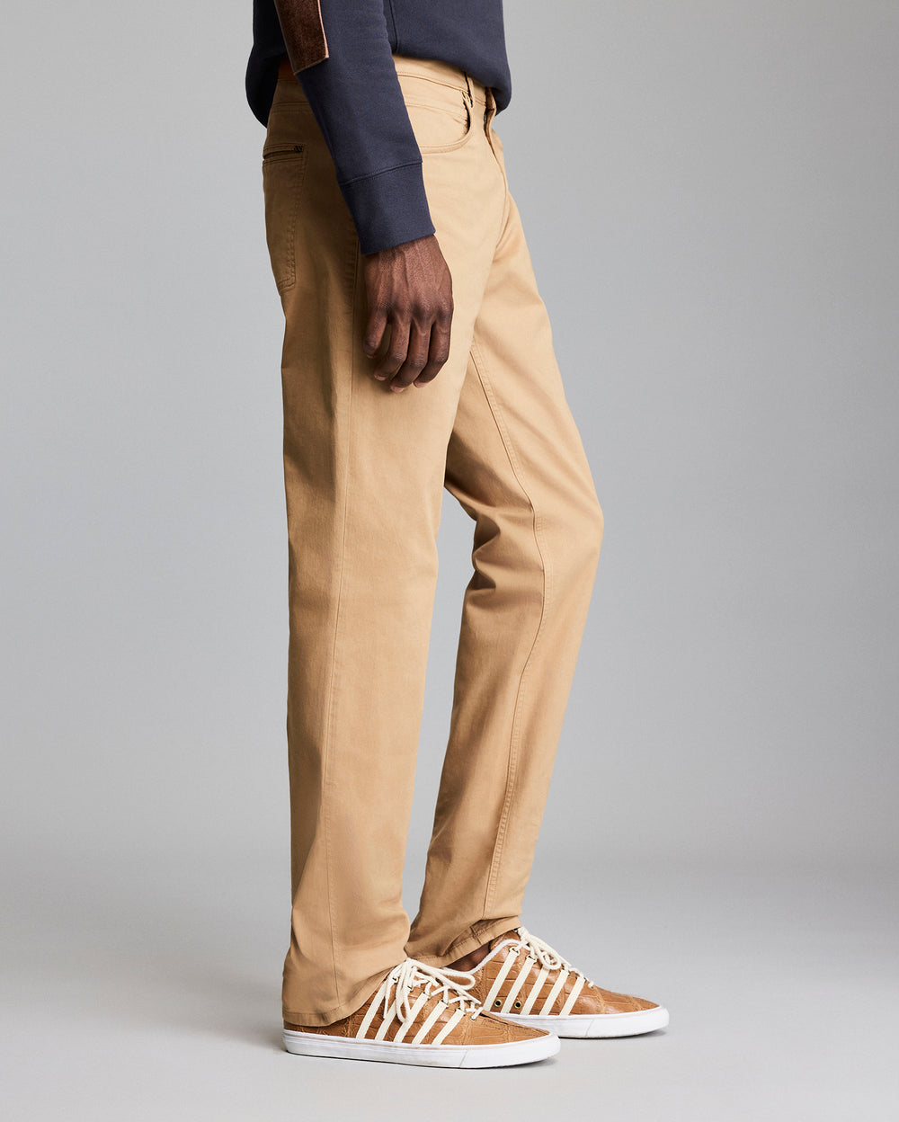A person wearing tan khaki pants, a dark long-sleeve shirt, and brown sneakers with white laces is standing against a plain gray background. Only the lower half of the body is visible.