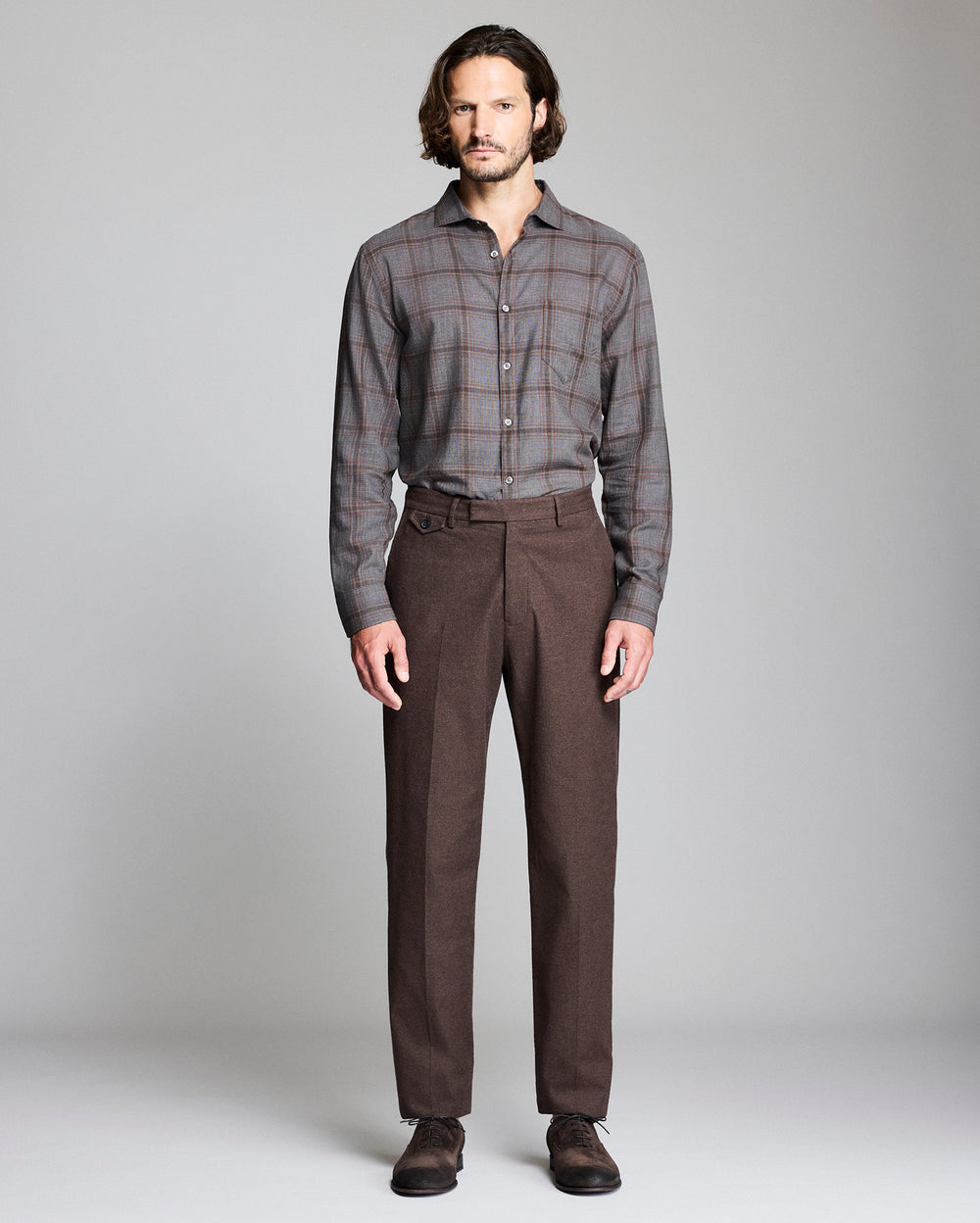 A man stands facing forward against a plain gray background, wearing a gray plaid button-up shirt tucked into brown dress pants, with dark brown shoes.