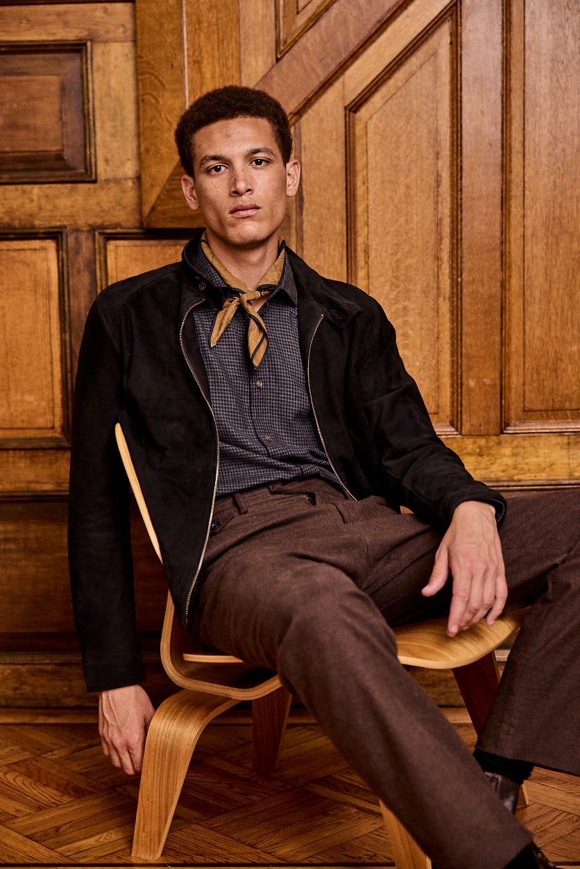 A man sits on a wooden chair in a room with wooden paneled walls. He wears brown pants, a checked shirt, a black jacket, and a light brown neck scarf, looking confidently at the camera.