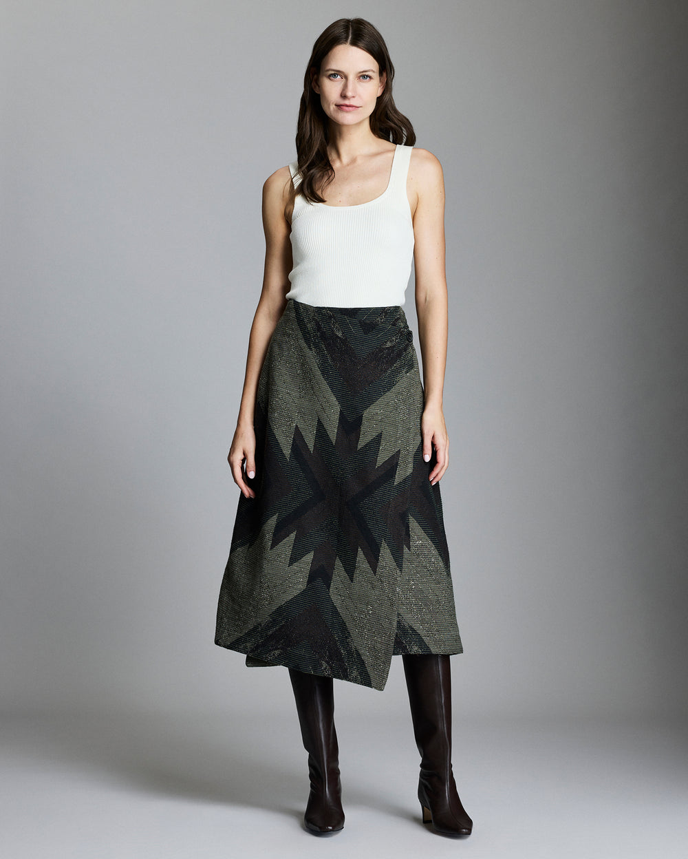A woman stands against a plain background, wearing a white sleeveless top, a dark patterned A-line skirt with a geometric design, and knee-high black boots. She has long brown hair and a neutral expression.