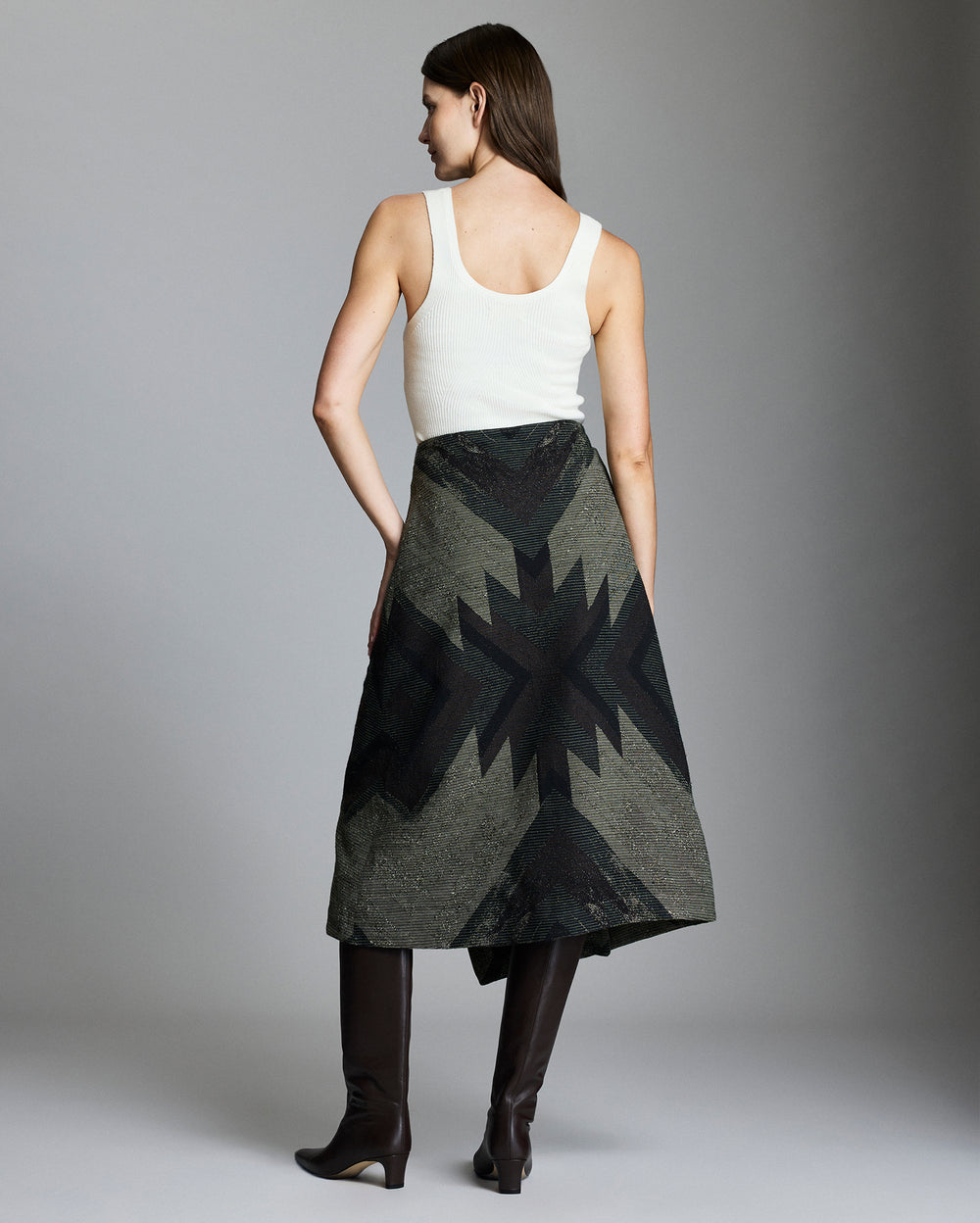 A woman stands facing away, wearing a white sleeveless top, an olive green skirt with a black geometric star pattern, and knee-high dark brown boots. The background is plain and gray.