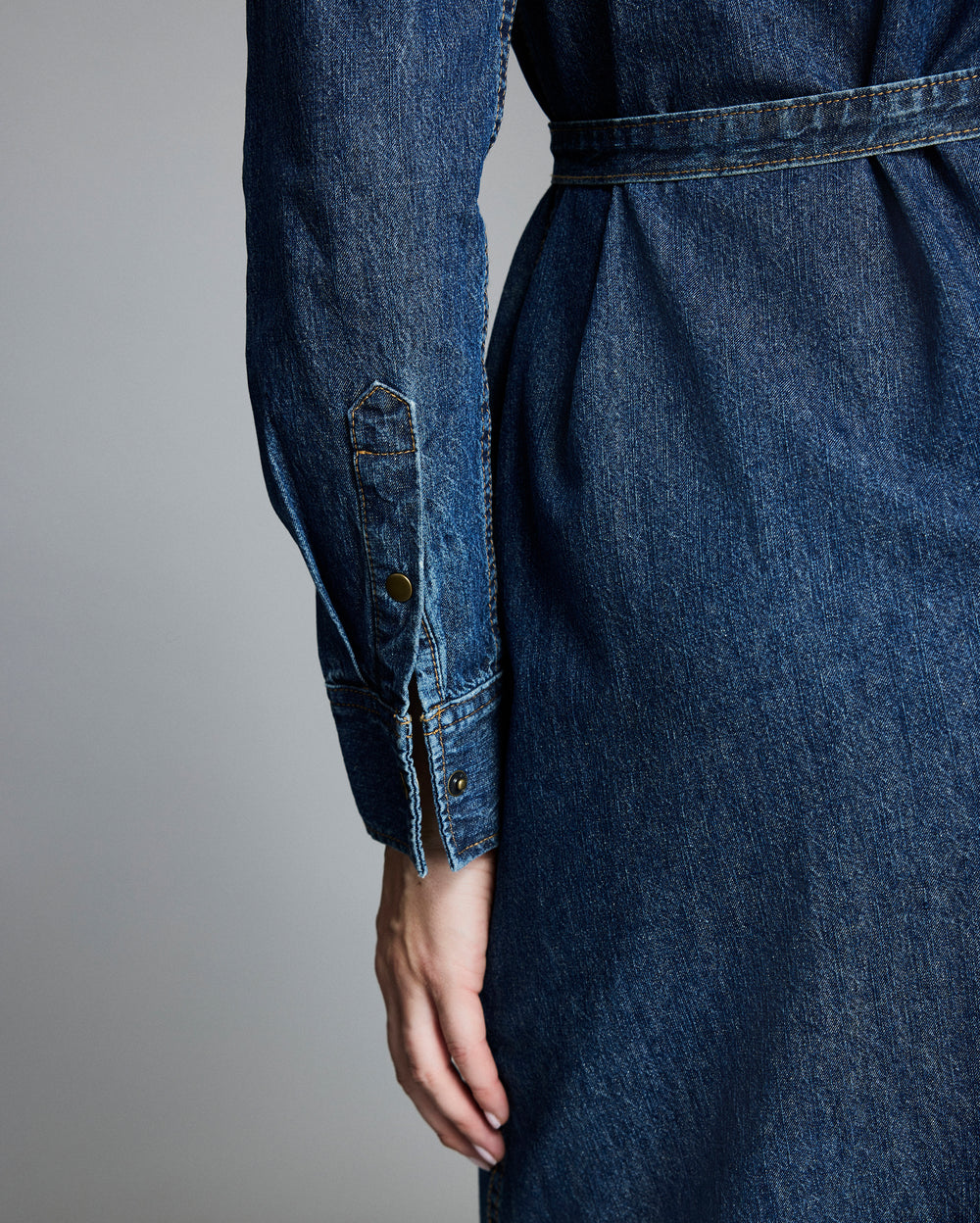 A close-up of a person’s hand resting at their side, wearing a long-sleeved, belted denim dress, shown from the back with details of the sleeve cuff and stitching visible.