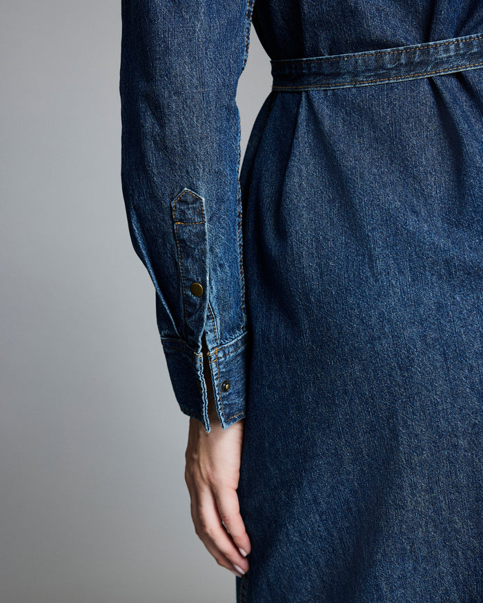 A close-up of a person’s hand resting at their side, wearing a long-sleeved, belted denim dress, shown from the back with details of the sleeve cuff and stitching visible.