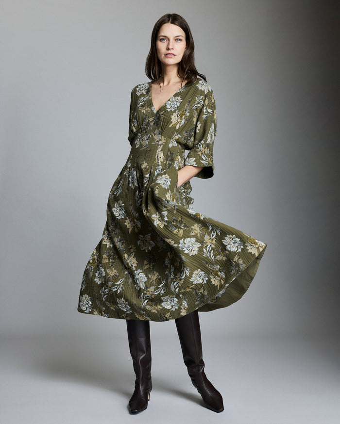 A woman stands in a studio wearing a long, olive-green floral dress with a v-neck and elbow-length sleeves, paired with tall dark brown boots. She has long brown hair and a neutral expression. The background is gray.