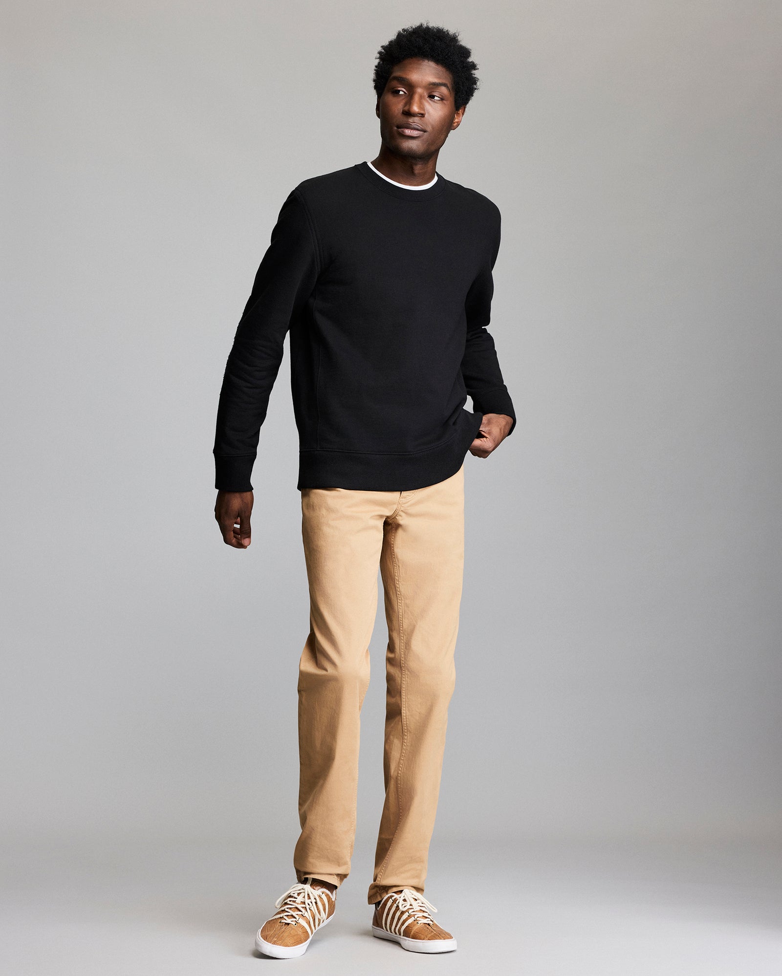 A man stands against a plain gray background, wearing a black long-sleeve shirt, tan pants, and tan sneakers with white laces, with one hand in his pocket.
