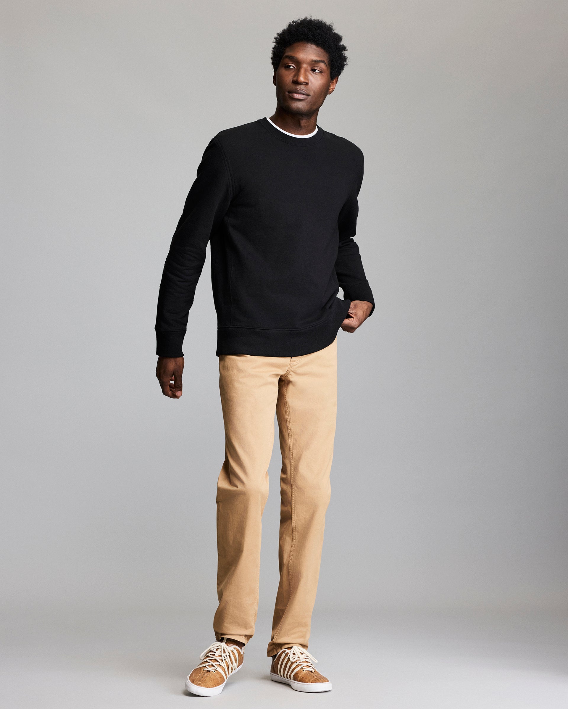 A man stands against a plain gray background, wearing a black long-sleeve shirt, tan pants, and tan sneakers with white laces, with one hand in his pocket.