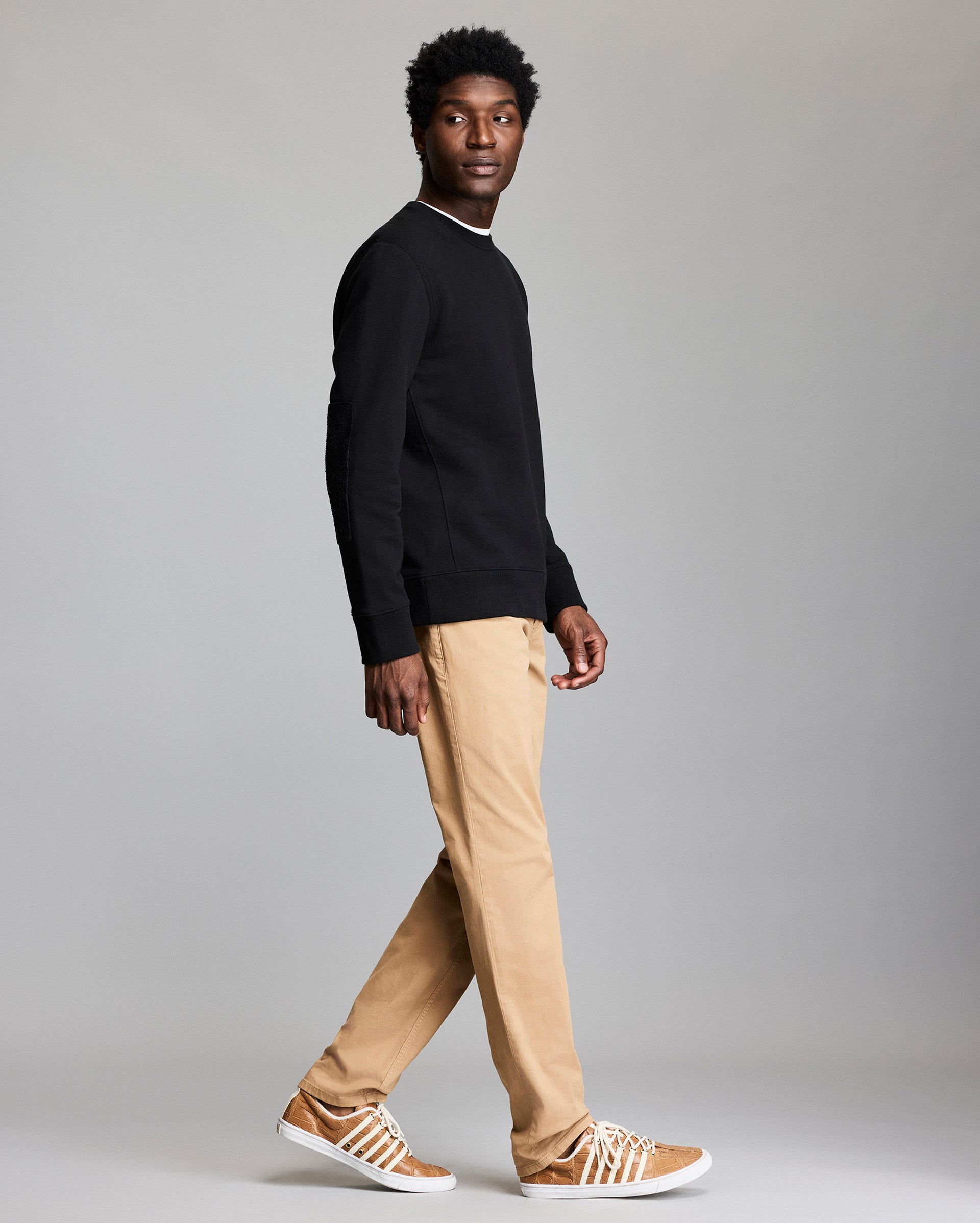 A man wearing a black sweatshirt, tan pants, and tan sneakers with white stripes walks sideways while looking slightly over his shoulder against a plain gray background.