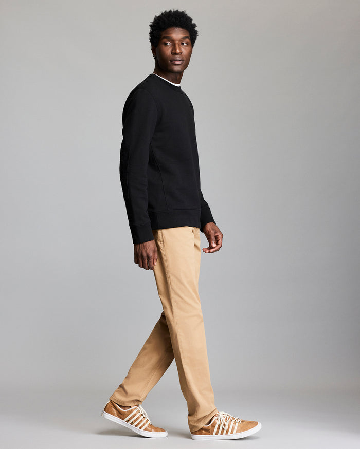 A man wearing a black sweatshirt, tan pants, and tan sneakers with white stripes walks sideways while looking slightly over his shoulder against a plain gray background.