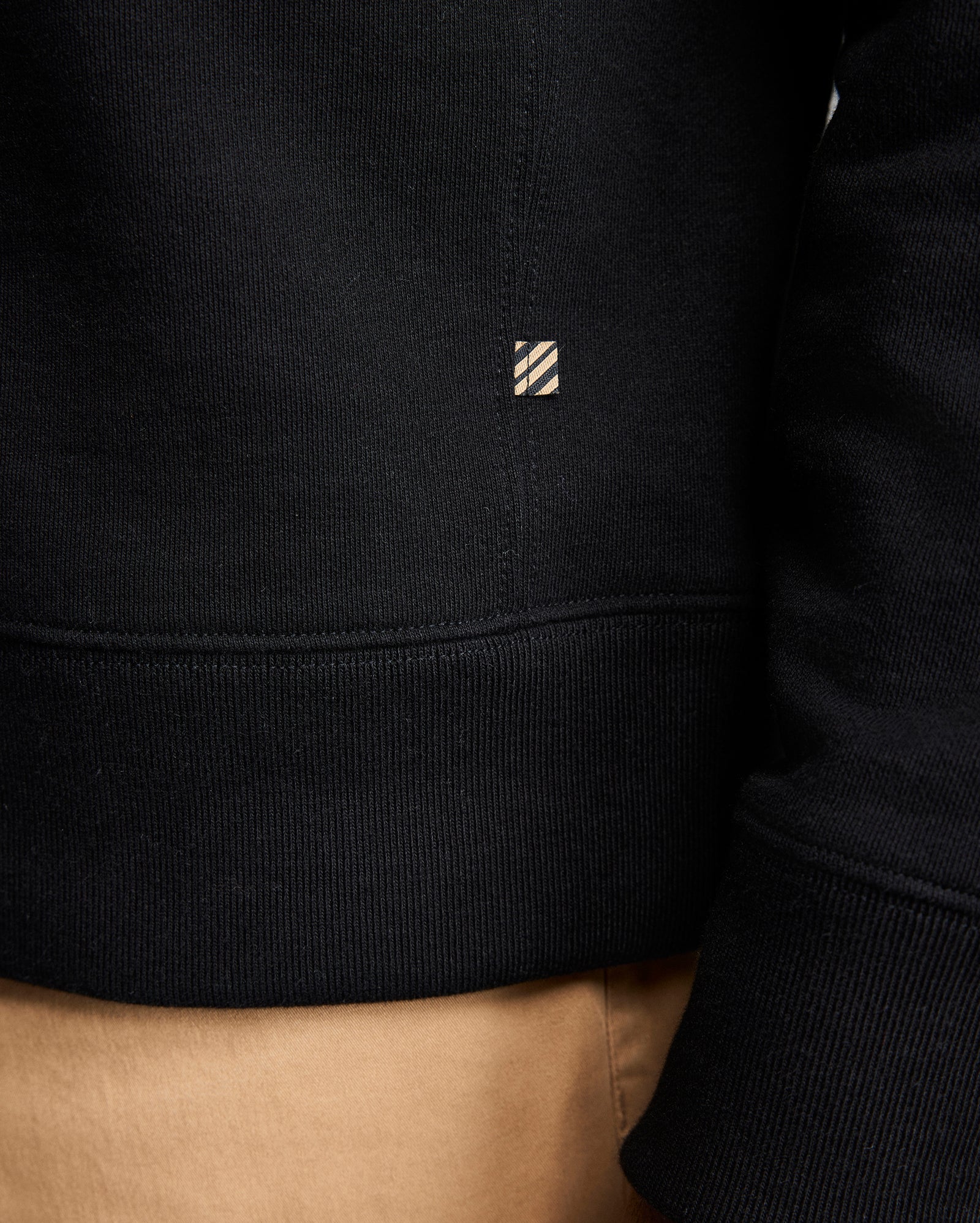 Close-up of a person wearing a black sweatshirt and tan pants. The sweatshirt features a small rectangular tag with diagonal stripes near the seam above the waistband. The persons arm and part of their side are visible.