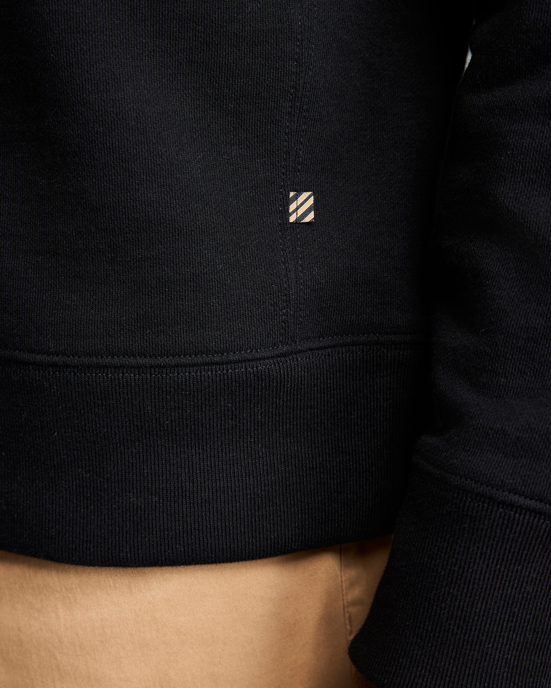 Close-up of a person wearing a black sweatshirt and tan pants. The sweatshirt features a small rectangular tag with diagonal stripes near the seam above the waistband. The persons arm and part of their side are visible.