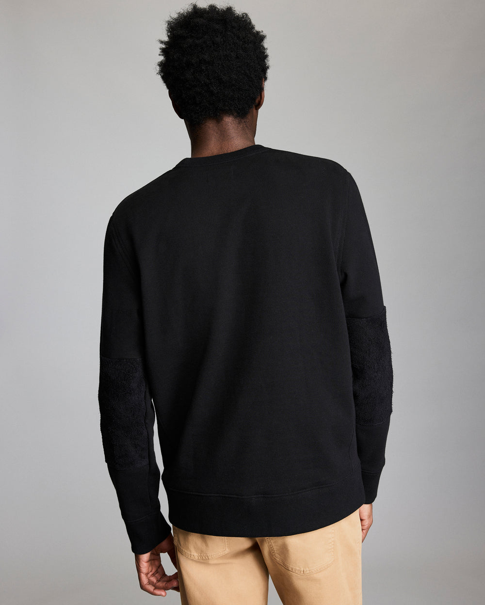 A person with short curly hair is standing with their back to the camera, wearing a black long-sleeve shirt and light tan pants against a plain, light gray background.