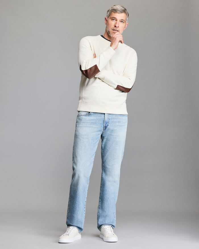 A man with gray hair and a beard stands thoughtfully against a plain background, wearing a cream sweater with brown elbow patches, light blue jeans, and white sneakers.