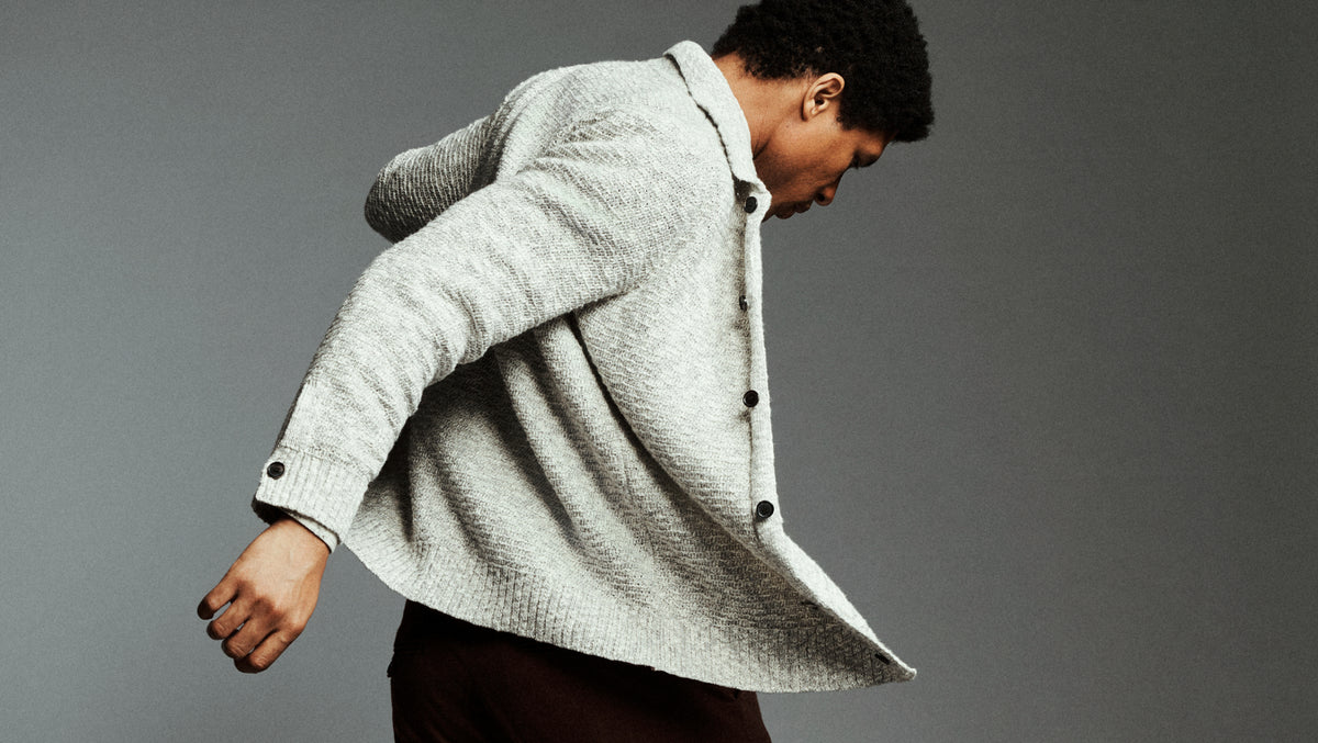 A man wearing a light gray textured knit cardigan sweater and dark brown trousers, captured in a dynamic pose against a solid gray studio background.
