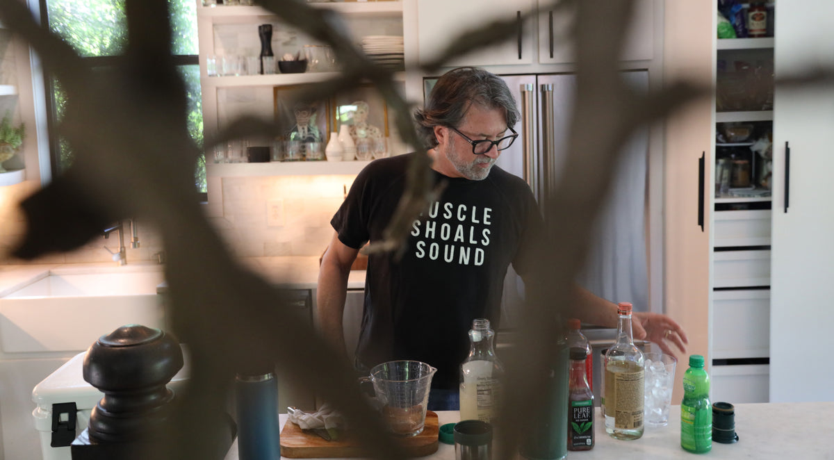 A man wearing a black t-shirt with the white text 'MUSCLE SHOALS SOUND' stands in a modern kitchen, reaching for items on a counter. The photo is viewed through a dark, blurry foreground branch or curtain.