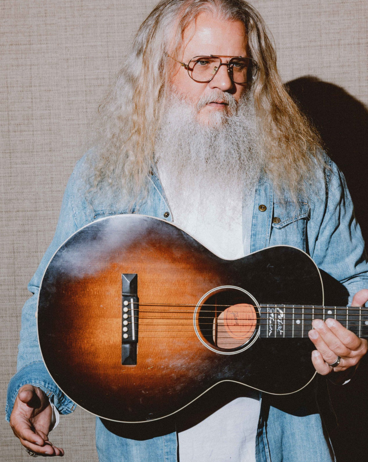 A man with long gray hair and a white beard, wearing glasses and a light blue denim shirt, holding a sunburst acoustic guitar against a textured beige background.