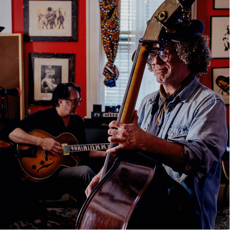 A man in a blue denim shirt and black hat plays a large double bass in a warmly lit room with red walls and framed pictures, while another musician sits in the background playing an electric guitar.