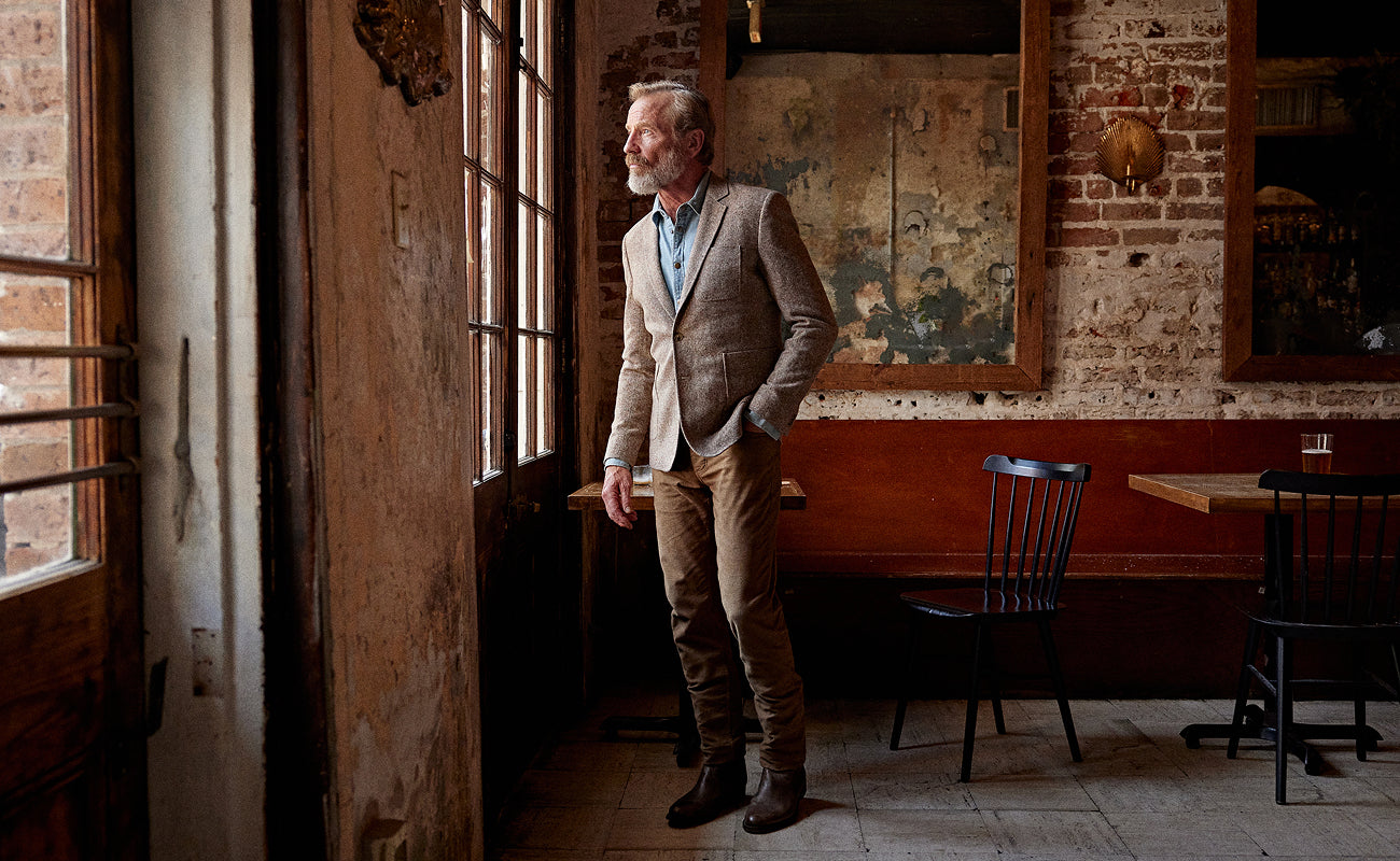 A man with a gray beard and glasses stands in a rustic, dimly lit room with brick walls and wooden doors, wearing a light brown tweed blazer, tan trousers, and dark brown boots.