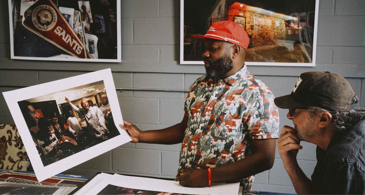 A man in a red cap and camouflage shirt holds up a framed photographic print showing a street scene, while another man in a dark cap looks on in a studio setting with framed art on a gray brick wall.