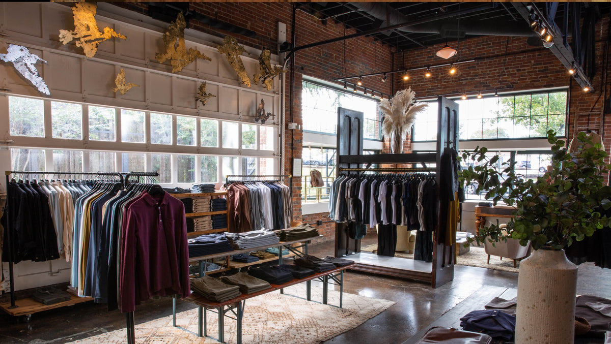 A clothing boutique interior with racks of apparel, tables with folded garments, brick walls, large windows, and decorative dried plants and wall art.