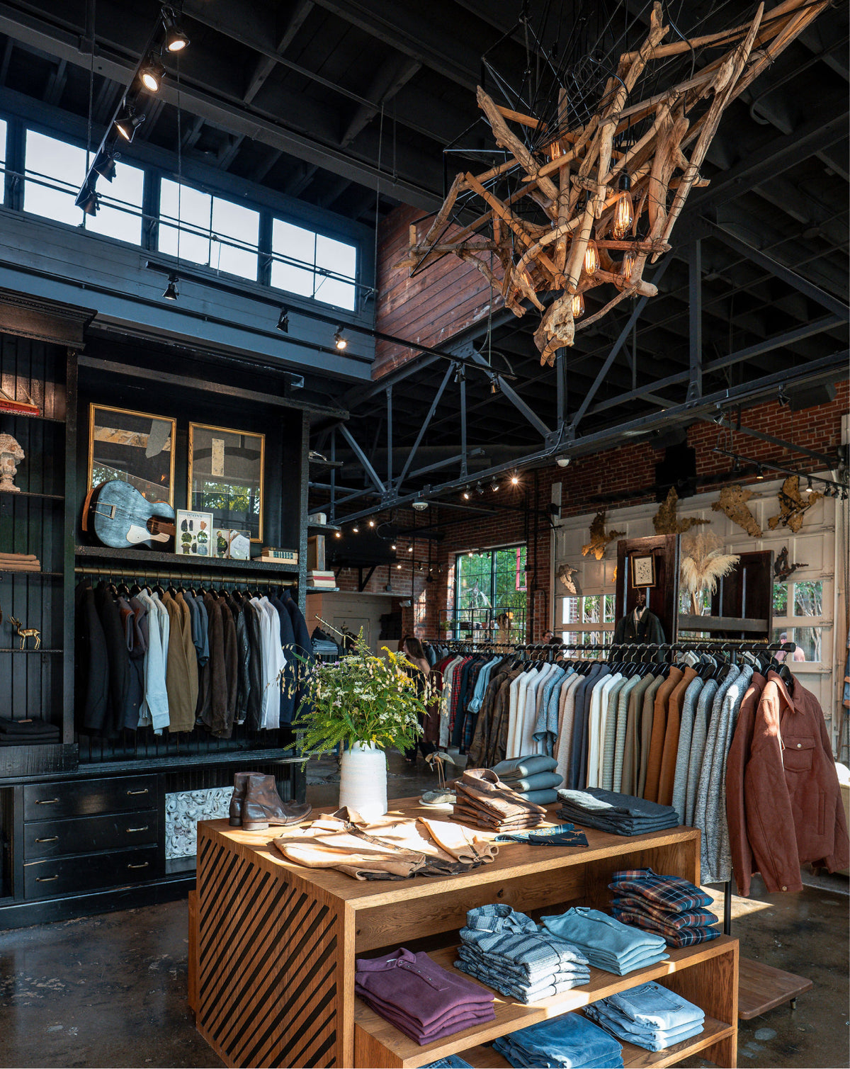 A rustic, industrial-style clothing boutique interior with clothing racks, a wooden display table with folded garments, a large natural wood branch chandelier, brick walls, and high windows.