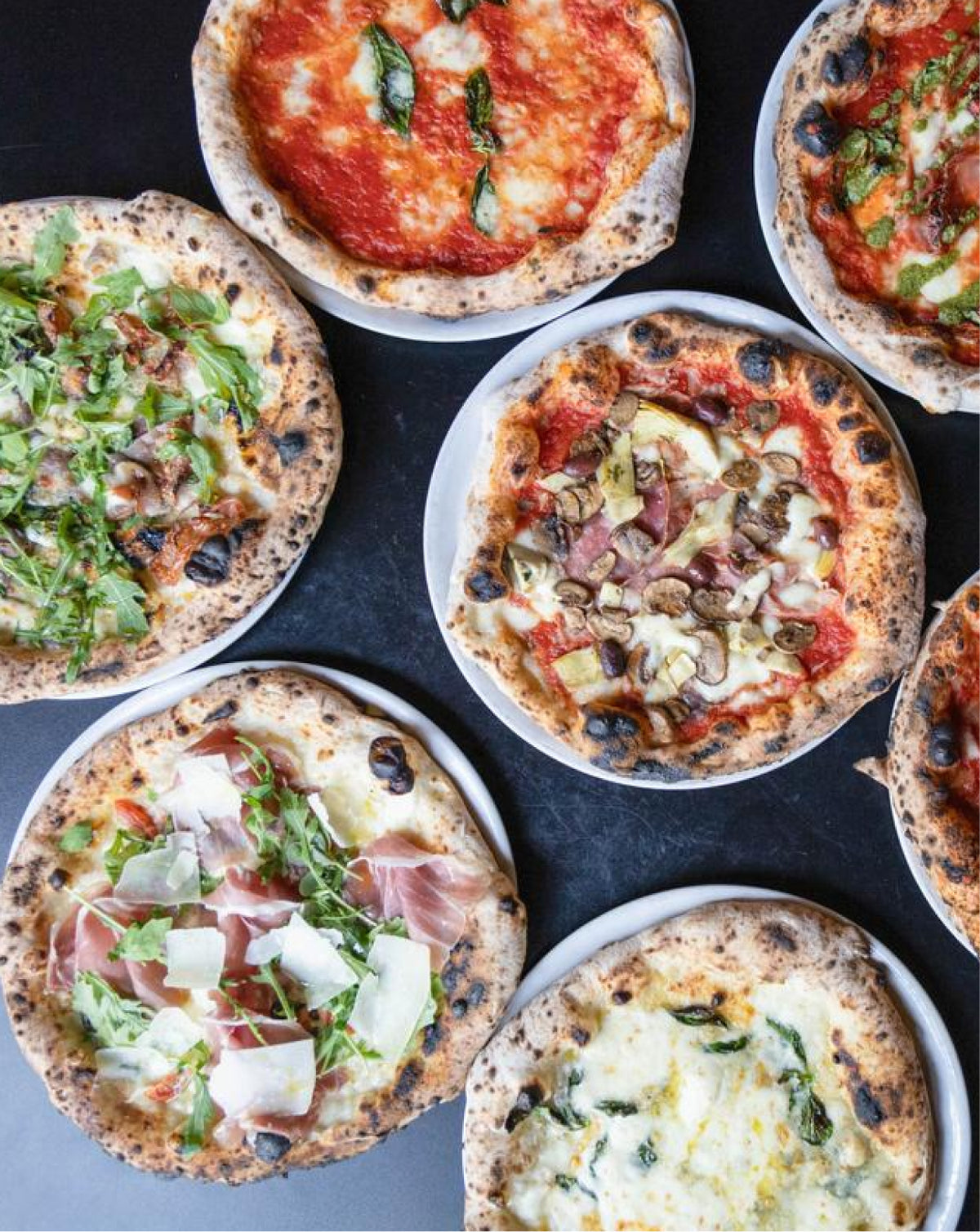 An overhead flat-lay view of several freshly baked artisanal pizza pies on white plates, arranged on a dark slate surface. The pizzas feature various toppings including fresh basil, prosciutto, arugula, mushrooms, and tomato sauce.