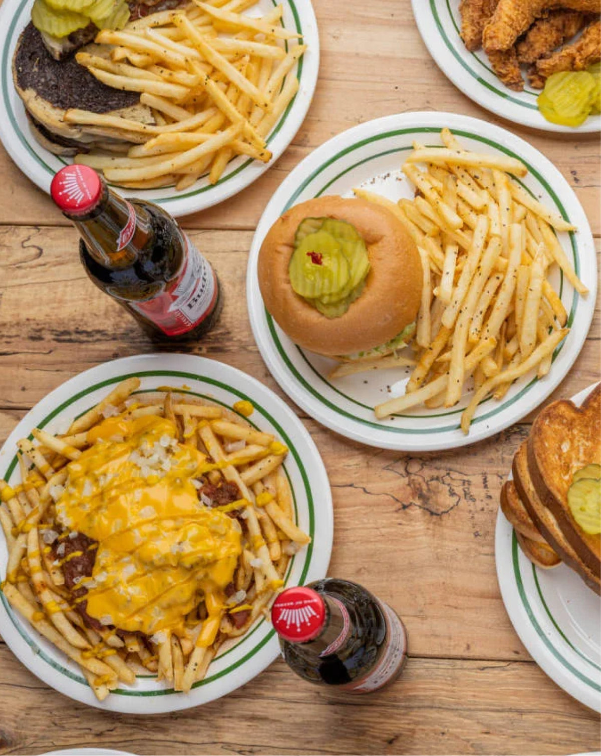Overhead view of a rustic wooden table set with multiple plates of food, including french fries, burgers topped with pickle slices, chili cheese fries, and two dark soda bottles with red caps.