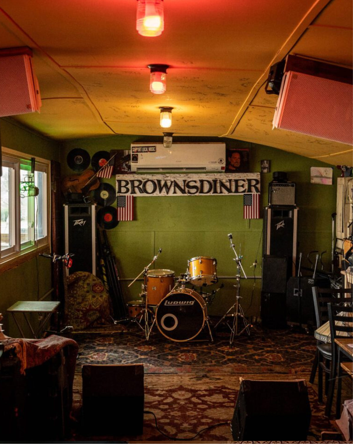 A vintage drum kit set up in a rustic, dimly lit mobile home or trailer with green walls, a 'BROWNSDINER' sign, and warm overhead lights casting a red and yellow glow.