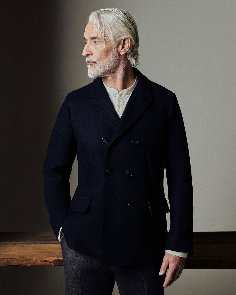 An older man with white hair and a beard wearing a dark navy double-breasted blazer over a cream shirt, standing in a studio setting.