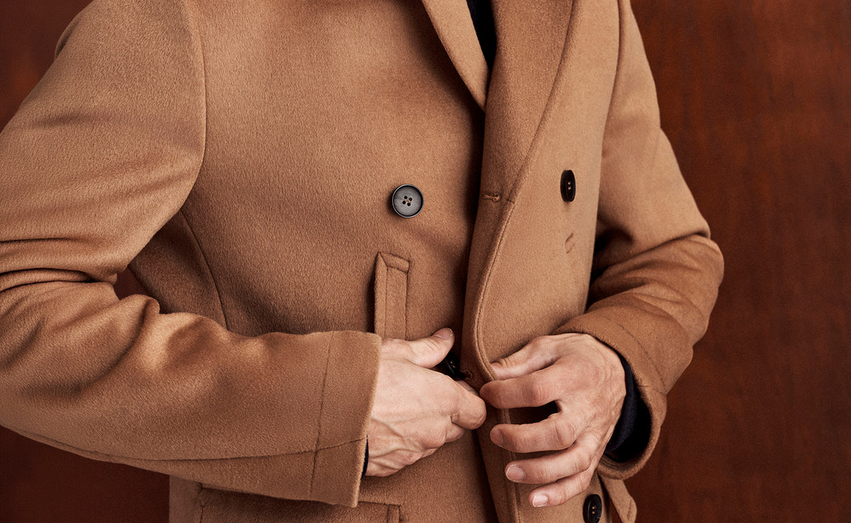 A close-up of a person wearing a camel brown double-breasted coat, adjusting the front flap with both hands against a dark brown background.