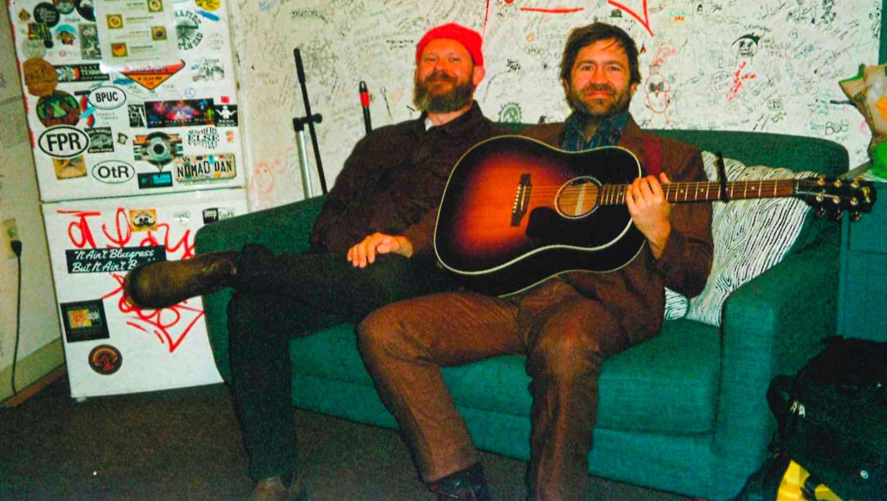 Two men sitting on a teal couch holding an acoustic guitar in a room with a sticker-covered refrigerator and a graffiti-style wall.