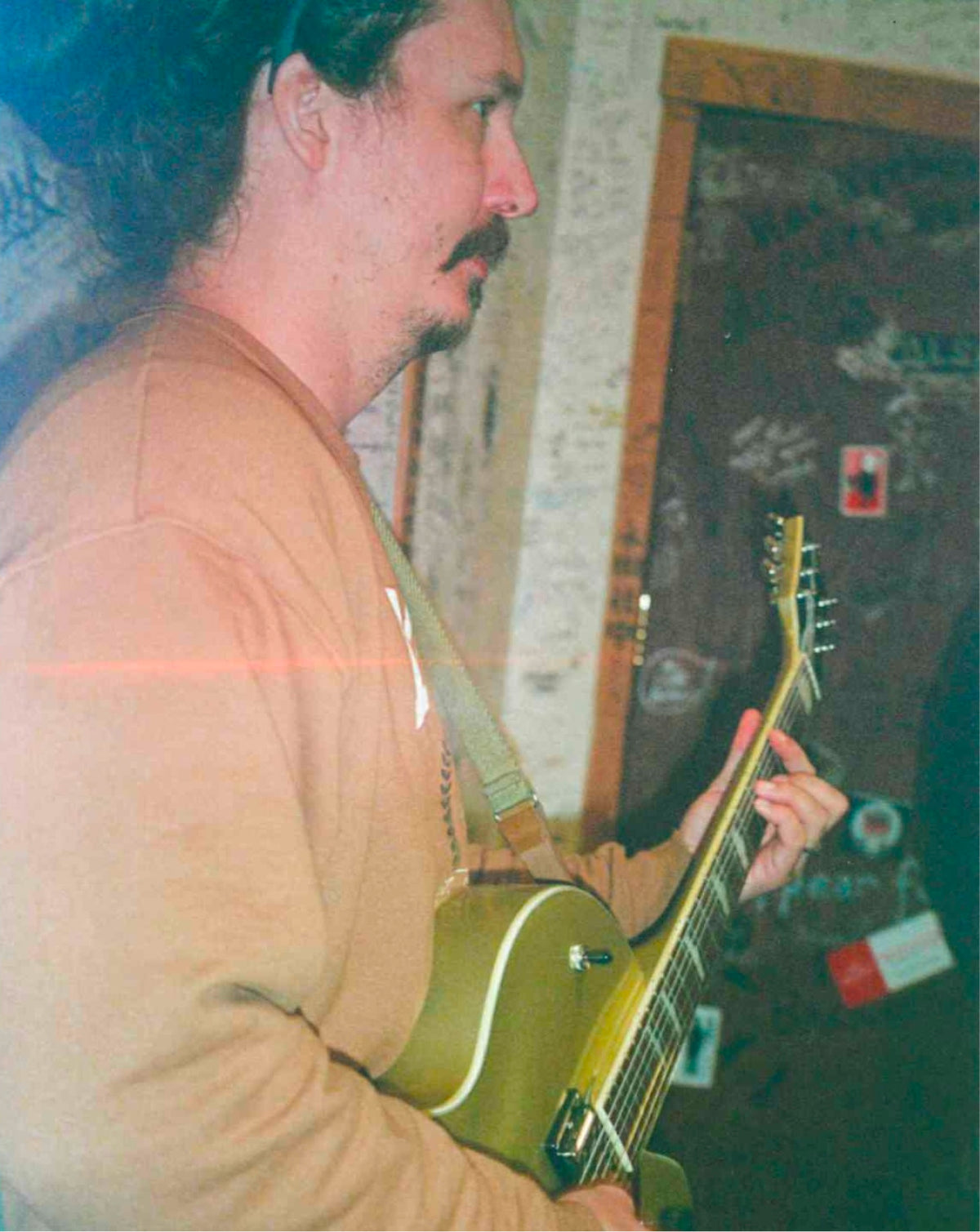 A man in a tan sweatshirt playing an olive green electric guitar in an indoor setting with a graffiti-covered door in the background.